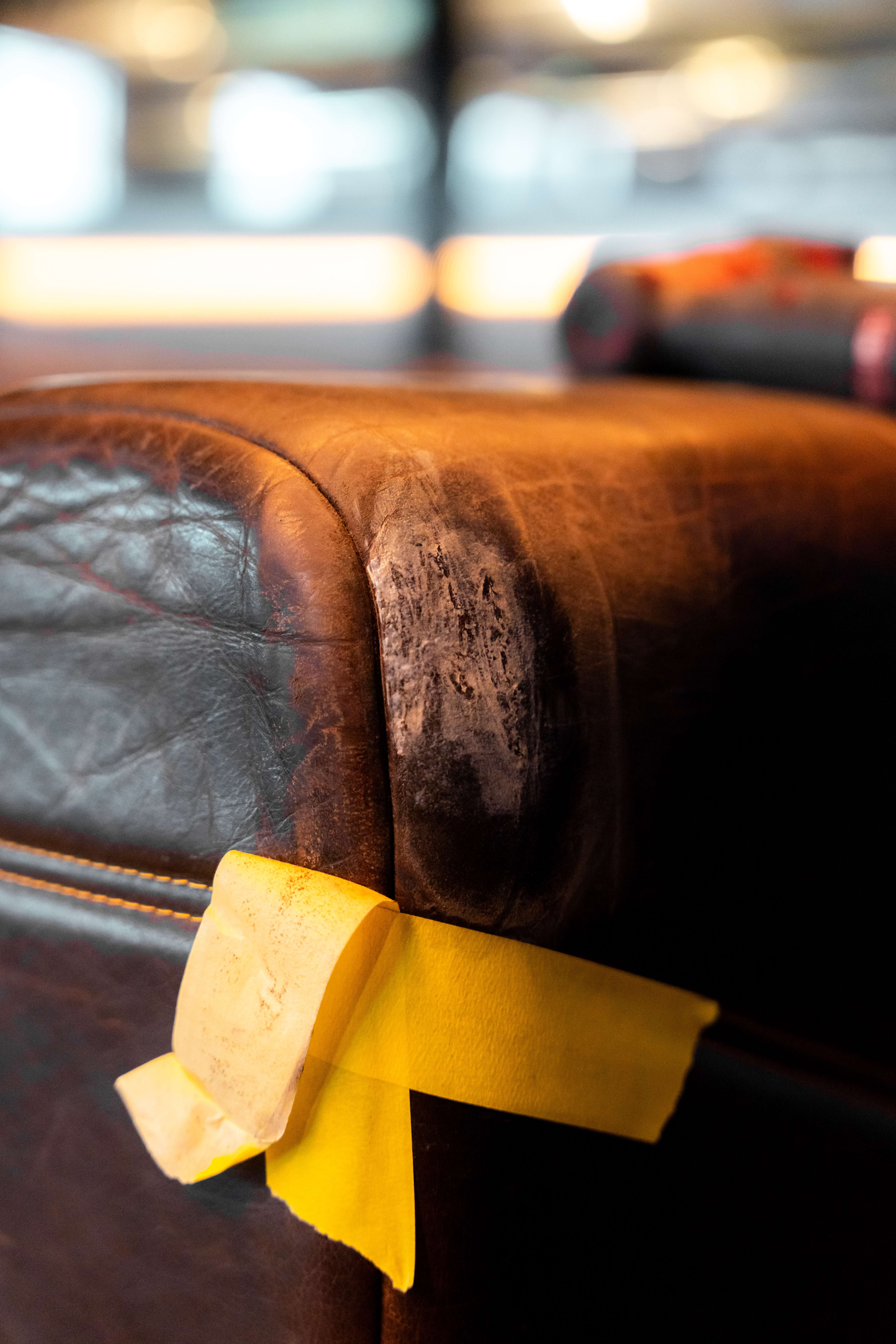 Dark brown leather banquette arm masked with yellow tape during an overnight shift, the worn corner prepared for repair with the venue lights on in the background