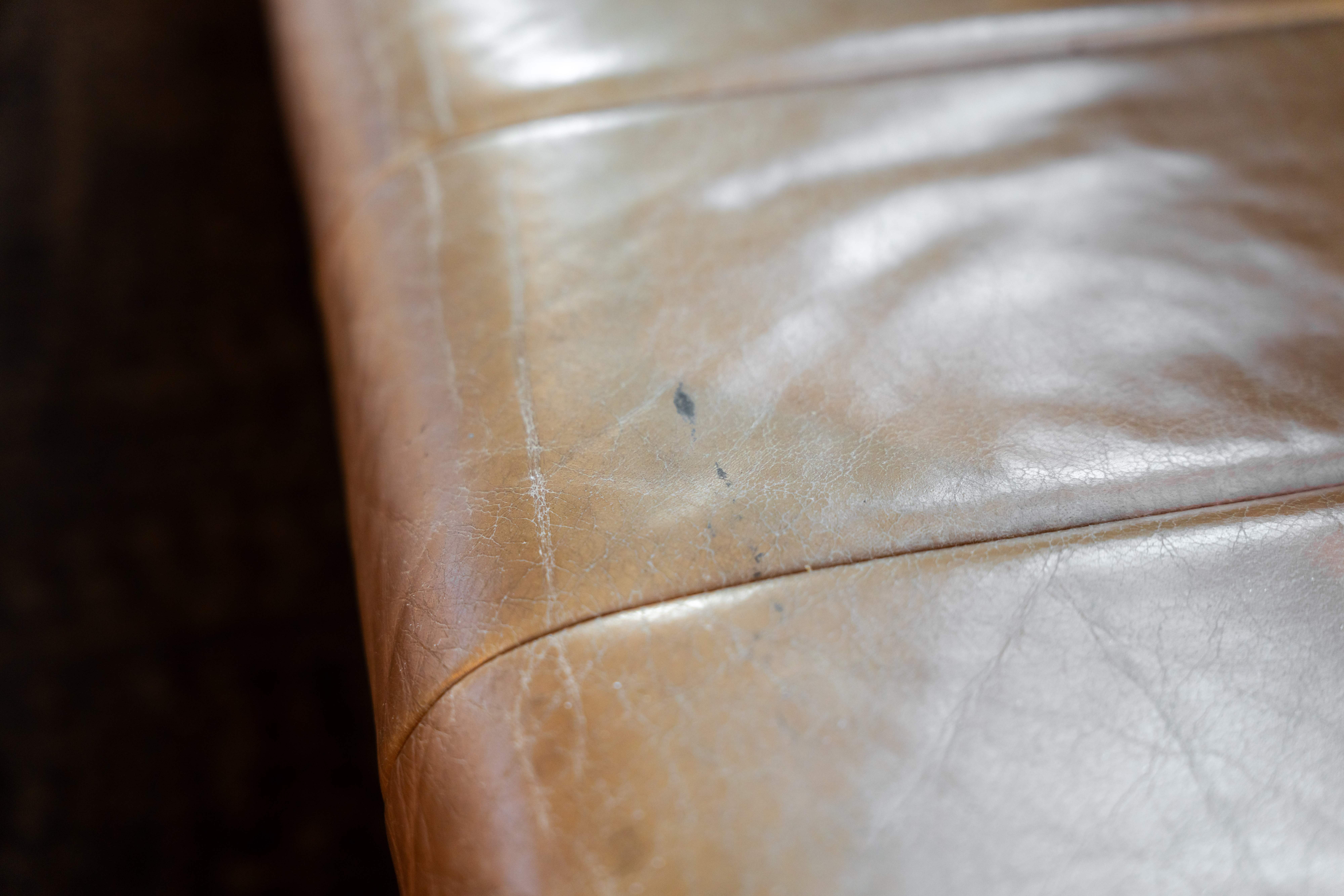Close up of a tan leather venue seat before restoration showing fine surface cracking along the seam and dark ink marks ground into the grain